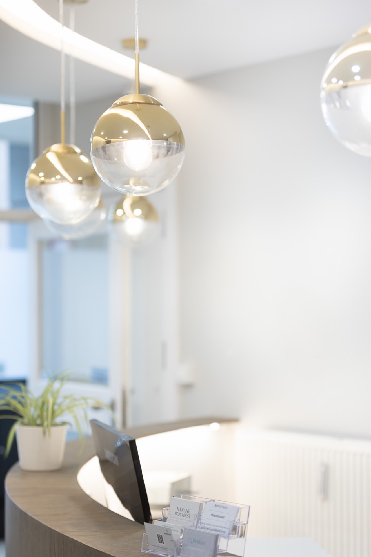 Medinoh — accueil cluster de globes laitons-pendants suspendus, comptoir chêne fumé courbe avec liseré LED, silhouettes en flou de mouvement
