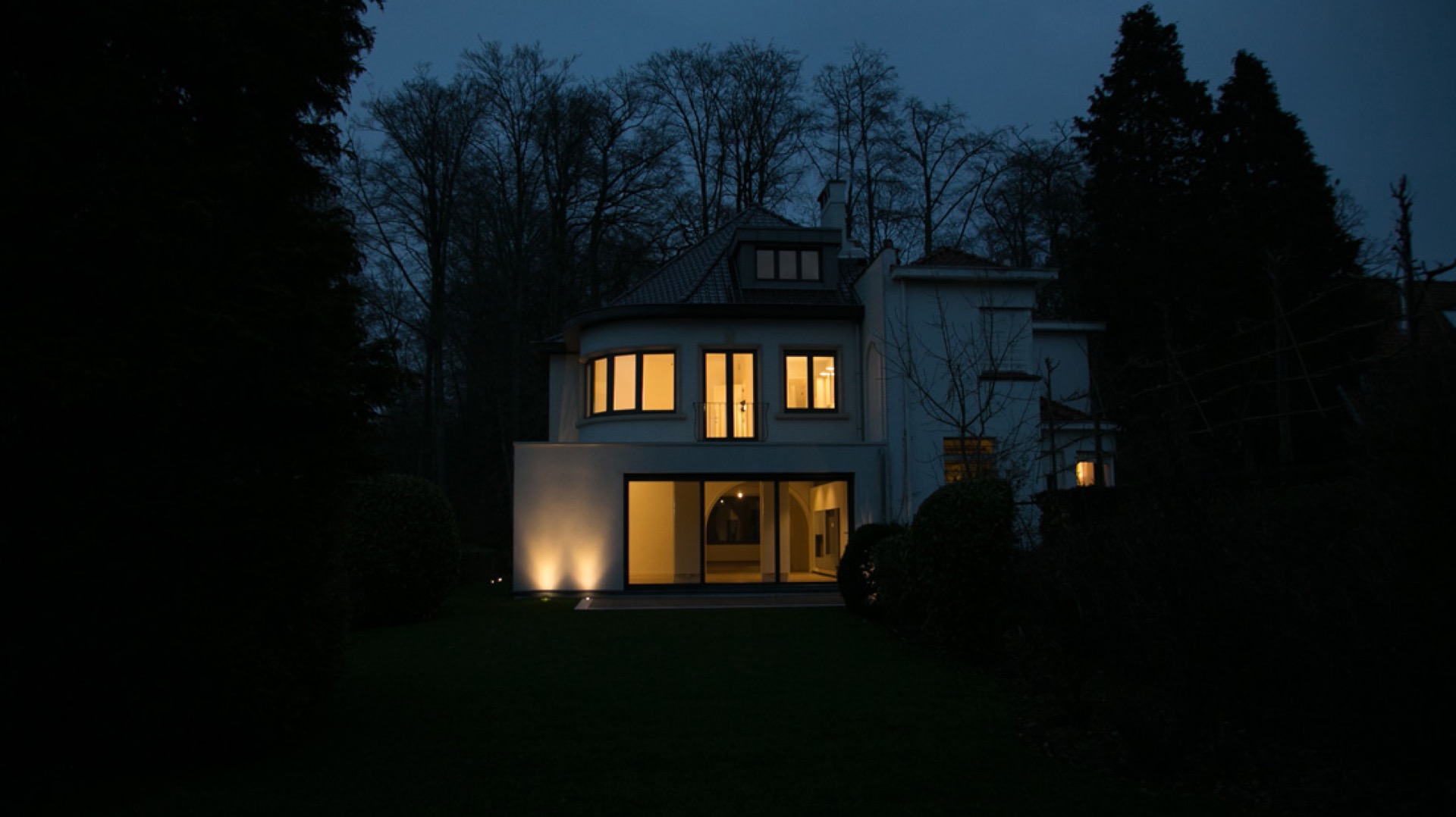 Statuaires 13 — villa blue hour vue jardin, façade arrière éclairée intérieur warm-light, baies vitrées rougeoyantes, masse arborée silhouettée